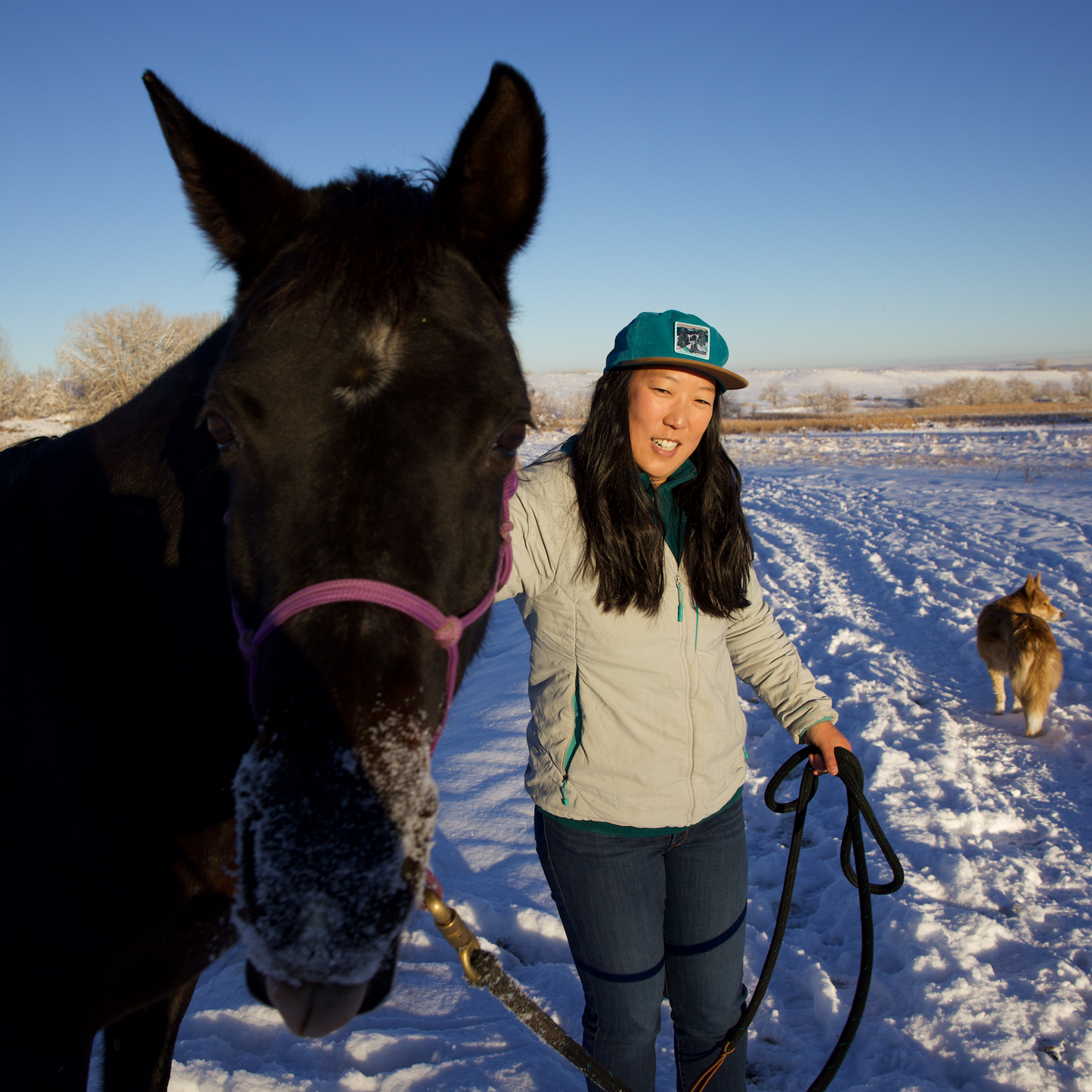 Woman standing with a horse and dog in a snowy field wearing Katherine Homes hat Winter Snowfall