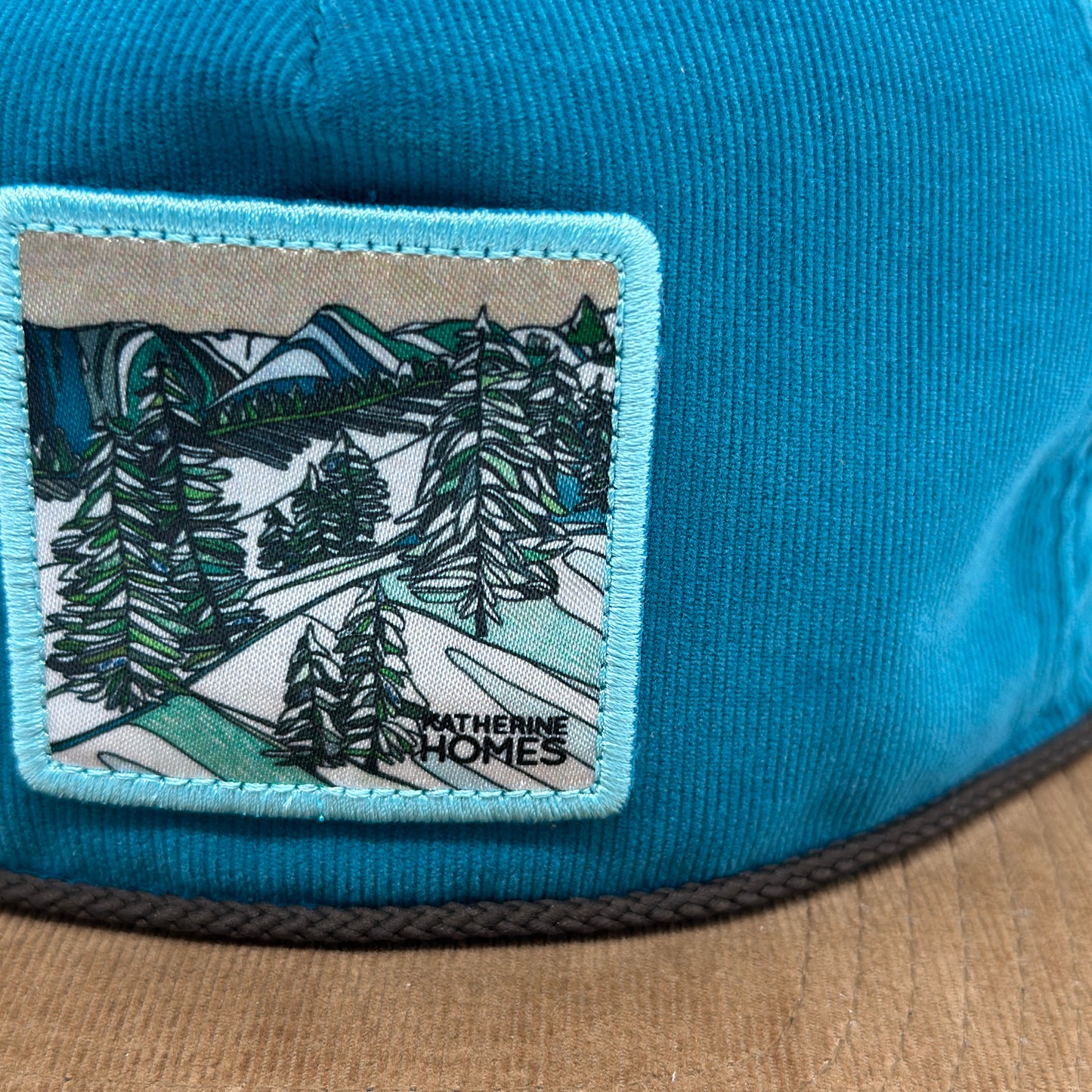 Blue cap with an embroidered patch featuring trees and mountains, Katherine Homes artist hat, Winter Snowfall